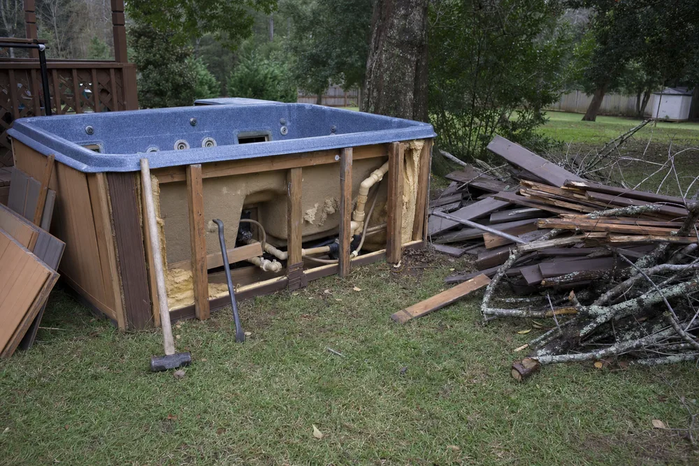An old backyard hot tub being dismantled next to a pile of cut deck boards and yard debris